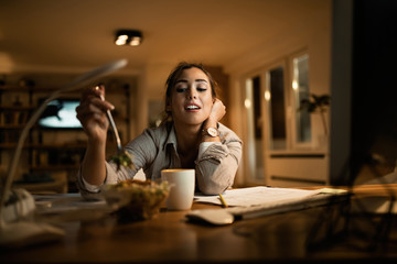Female student eating salad while studying at night at home.