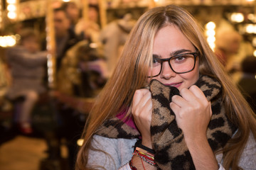 Fototapeta premium Blonde girl in cold, warms up with scarf on a Christmas night. Carousel in the background