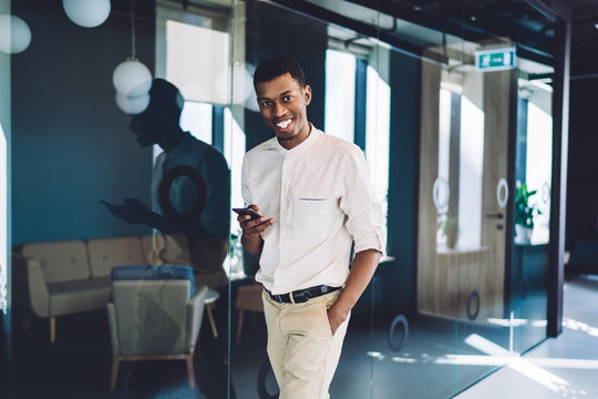 Cheerful ethnic businessman texting smartphone and walking along office hall