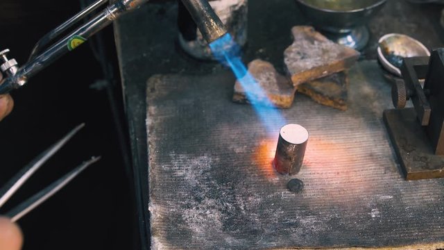 CU shot: skilled worker heats cylindrical metal bar with hot gas burner on large asbestos sheet in dark workshop closeup