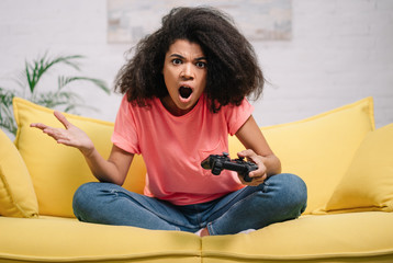 Emotional woman playing video games, sitting on yellow sofa holding wireless joystick. Excited African American hipster girl with wide open mouth lose game © Oleksii