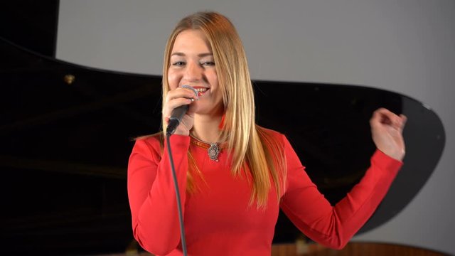 A girl sings into a microphone against a piano background on stage in a red dress