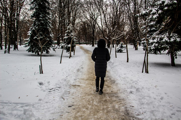 A girl in a black coat is walking along the road. Silhouette of a girl. Winter road. The girl in the hood in the winter. Black clothes. A winter day with trees and snow.
