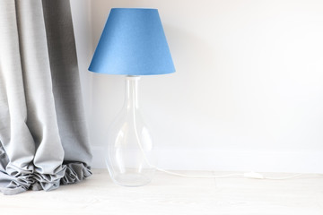 Blue lampshade on the table lamp with glass element near the light wall and grey cloth. Trendy color in your interior. Grey and blue colors.