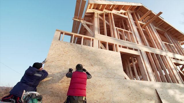 Two professional builders are covering the wooden house frame with plywood using hydraulic hammer. Wooden frame house under construction