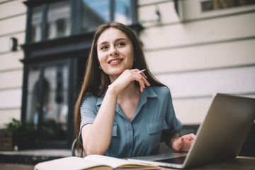 Young female freelancer in casual wear using laptop and making notes at cafe