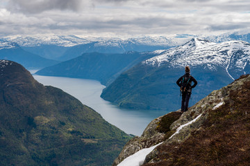 Fototapeta na wymiar An explorer stands atop a mountain looking across a sweeping view of Norwegian fjords and snow capped mountains