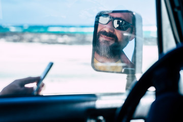 Portrat of man face reflected and view inside a car mirror -  people enjoying travel and adventure driving vehicle in outdoor scenic place - beach and sea water in background