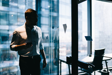 Young intelligent male employee with cellular device standing in office interior of corporate enterprise and thinking on strategy idea, formally dressed professional with smartphone technology