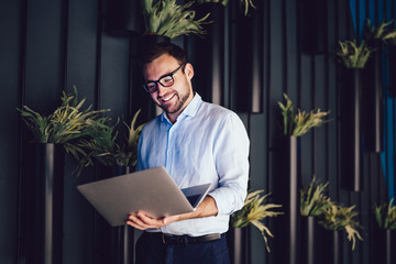 Cheerful marketer in optical spectacles reading email with good news about corporate analytic using...