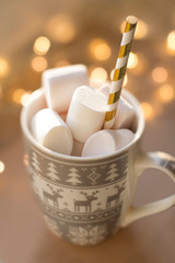 white christmas mug with reindeer design full of white marshmallows with gold straw with gold lights on beige background