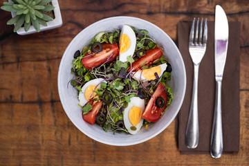 Fresh vegetable salad with mixed greens (arugula, mesclun, lettuce), eggs, black olives, tomato and bean sprouts. Wooden background with cutlery and a cactus. Top view