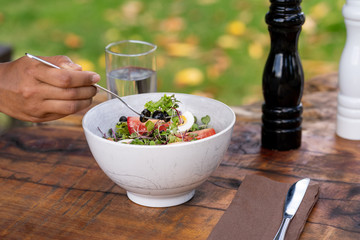 Hand pricking with a fork a salad with mixed green leaves, eggs, black olives and tomato on a wooden rustic table with a glass of water and pepper and salt shakers behind. Natural environment