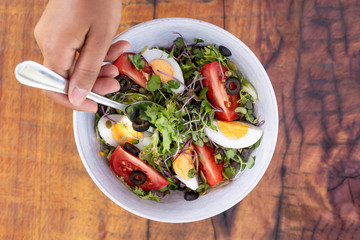 Hand pricking with a fork a salad with mixed green leaves, eggs, black olives and tomato on a wooden rustic table. Close up. Top view