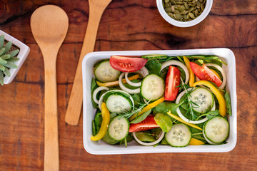 Fresh salad with cucumber, baby spinach, rosemary, onion, pumpkin seeds and tomato served on a square white plate and wooden cutlery. Wooden background. Top view