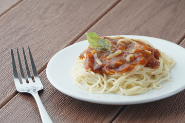 View of spagetti bolognese with green basil on top over brown background. Silver fork placed next to the white plate.