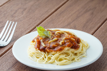 View of spagetti bolognese with green basil on top over brown background. Silver fork placed next to the white plate.
