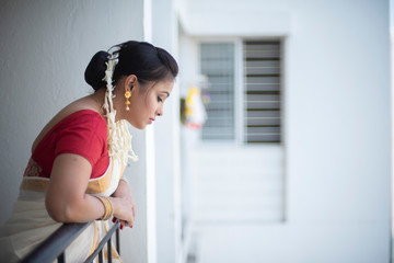 An young and attractive Indian woman in white traditional sari and red blouse and flowers is standing in a balcony for the celebration of Onam/Pongal. Indian lifestyle.