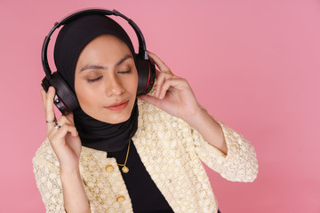 Beautiful young hipster girl wearing hijab and listening to music with wireless headphones isolated over pink background. Technology in a modern lifestyle concept.