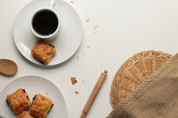 Flat lay of fresh, relaxed setting of black coffee and Pan Au Chocolate French inspired afternoon tea arrangement with clean white background with copy space and timber and brown accents and textures