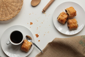 Flat lay of fresh, relaxed setting of black coffee and Pan Au Chocolate French inspired afternoon tea arrangement with clean white background with copy space and timber and brown accents and textures