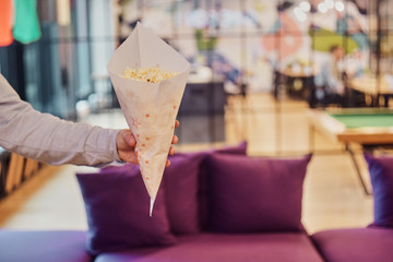 A man holds a paper bag with popcorn on the background of the office, hostel, co-working, restaurant, cinema. A man hands out  popcorn.