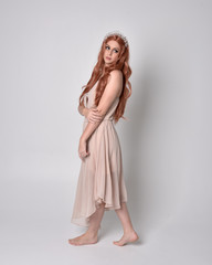 full length portrait of a pretty, fairy girl wearing a nude flowy dress and crystal crown. Standing and dancing pose against a grey studio background.