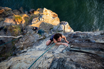 Adventurous Girl is Rock Climbing up a Steep Cliff during a summer sunset. Taken in Lighthouse Park, West Vancouver, British Columbia, Canada.