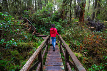 Woman wearing a red coat walking on a wooden path in a wild forest. Taken in Rainforest Trail, near Tofino and Ucluelet, Vancouver Island, BC, Canada.