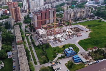 Naklejka premium Aerial view of the construction site of a building in the industrial center of the city among buildings located close to each other with three yellow high elevating cranes by a green lawn with grass