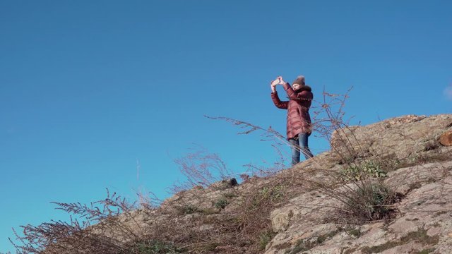 A young female tourist climbed to the top of the mountain. She stands on the edge of a cliff against a blue sky and takes pictures of the landscape on a smartphone.