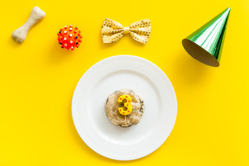 Birthday party for puppy concept. Dog feed, candle, bone, bow-tie and party hat on yellow background top-down