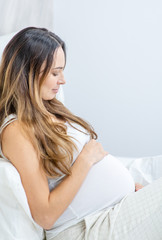 Naklejka premium happy pregnant woman sitting on bed and touching her belly at home