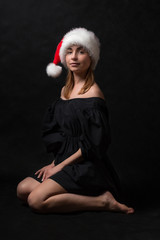 Fototapeta na wymiar A girl in a black dress and Santa Claus hat is sitting on the floor on a dark background.