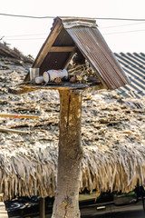 Old wooden spirit house with rusty roof stands neglected in the sun with a palm-thatched roof house in the background.