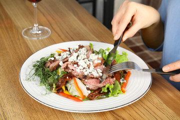 Woman eating delicious salad with roasted meat at wooden table, closeup