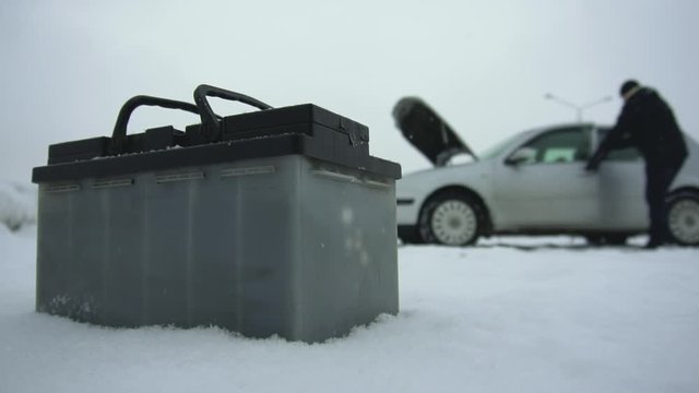 Man repairs or fixes broken car in winter snowfall. Malfunction or problem with auto. Automotive battery lying next to the vehicle. Hood or bonnet raised up. Typical lead-acid battery is on foreground