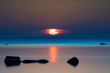 Vibrant colored summer sunset reflecting in ocean with endless horizon and deep blue ocean, silhouette of boulders laying in the foreground in shallow water at island of Gotland, Sweden