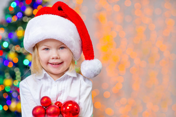 little girl in a Christmas hat holding christmas balls against the background of holiday lights