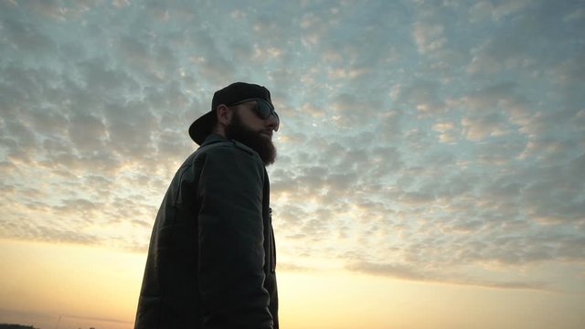 Handsome stylish caucasian guy model in black glasses with a beard looks into the distance against the backdrop of a sunny sunset in the city, background. Slow motion
