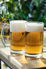 Subject shot of classic design beer mugs with frothy pale amber ale. The glass mugs with thick handles are isolated on the white wood table in the summer outdoor interior against glass bottles. 