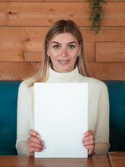 Cheerful blonde female model in a cozy warm cafe holds a white magazine front view with mockup blank cover