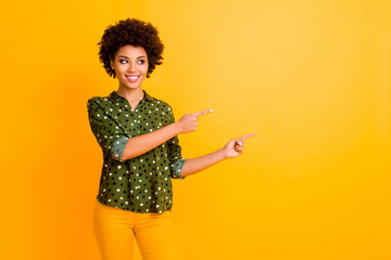 Photo of amazing dark skin curly lady indicating finger empty space advising buyer new product wear trendy green dotted shirt pants isolated yellow color background