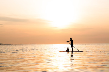 Fototapeta na wymiar Silhouette of dad and son playing the stand-up paddle board on the sea with beautiful summer sunset colors. Happy family concept.