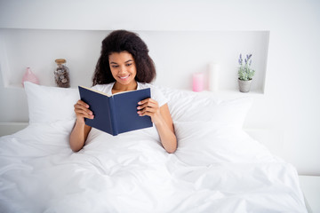 Photo of charming curly dark skin lady lying sheets bed covered white blanket hold book reading...