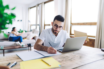 Fototapeta premium Adult man writing in notepad while sitting at laptop at home