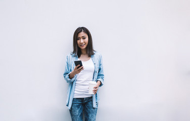 Young cheerful Asian woman leaning on wall with smartphone and takeaway coffee