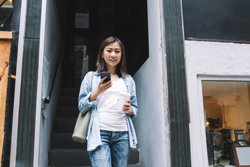 Obraz premium Cheerful female with mobile drinking takeaway coffee on stairs of shop on street