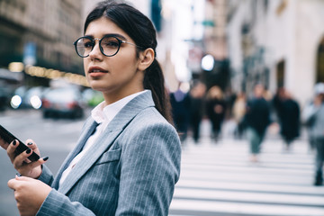 Pensive female entrepreneur standing at urban setting in downtown and thinking on received cellphone email with strategy information, contemplative Latino woman using smartphone gadget outdoors