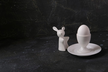 Boiled egg in a ceramic white stand on a saucer and a rabbit-shaped salt shaker on a dark background under the concrete. Breakfast. Minimalism.